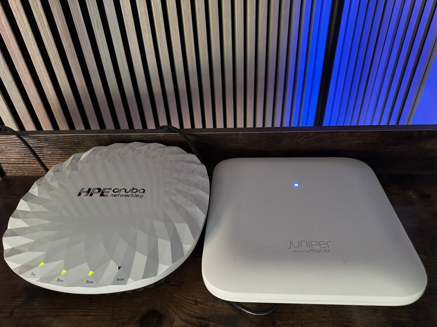 An HPE Aruba AP-735 and a Juniper AP34 lying side by side on a wooden shelf, both flat on their backs with their radomes facing up and status lights lit